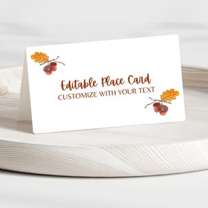 May include: White place card with the text "Editable Place Card CUSTOMIZE WITH YOUR TEXT" in brown. The card is decorated with watercolor illustrations of acorns and fall leaves. The card is displayed on a wooden tray.