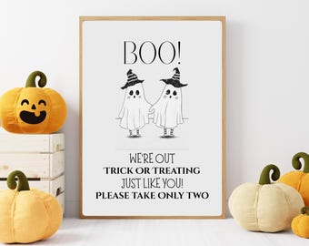 May include: A framed Halloween sign with the word "BOO!" at the top and two ghost illustrations wearing witch hats. The sign also includes the text "WE'RE OUT TRICK OR TREATING JUST LIKE YOU! PLEASE TAKE ONLY TWO". Decorative plush pumpkins are in the foreground.