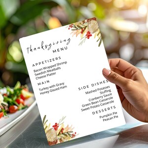 May include: A Thanksgiving menu card held by a person. The menu features appetizers like bacon-wrapped shrimp and Swedish meatballs, main courses such as turkey with gravy, and side dishes including mashed potatoes. Floral accents decorate the corners.