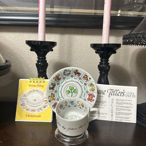 Op de afbeelding: Een waarzeggerij theeservies met een schotel met dierenriemtekens en een klaverblad. De theekop heeft de tekst "Ask it thy fortune" en staat op een heldere standaard. Een gele kaart met "Fortune Telling" en "TEACUP" is ook aanwezig.
