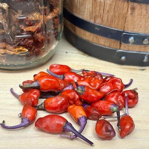 May include: A pile of small, red chili peppers with purple stems. The peppers are arranged on a light brown wooden surface.