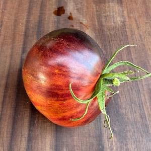 May include: A ripe, heirloom tomato with a deep red and purple color, and green stem. The tomato has a round shape and is sitting on a wooden surface.