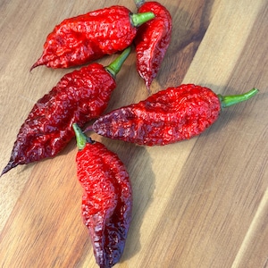 May include: Five vibrant red ghost peppers with textured skin and green stems are arranged on a wooden surface. The peppers have a deep red color that fades to a darker shade at the tips. The peppers are a popular ingredient in spicy dishes.