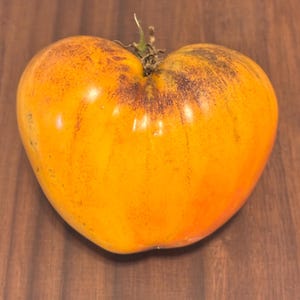 May include: A large, heart-shaped heirloom tomato with a vibrant orange hue, accented by darker areas near the stem. The tomato's skin has subtle variations in color, and a small green stem is visible at the top. The tomato rests on a wooden surface.