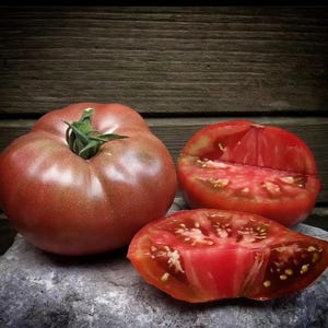 May include: Three heirloom tomatoes, one whole and two sliced in half, showing the red flesh and dark seeds. The tomatoes are on a gray stone surface.