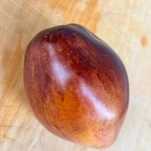 May include: A single, ripe plum with a deep burgundy skin and streaks of reddish-brown. The fruit has a smooth, glossy surface and is set against a light-colored wooden cutting board. The plum's shape is oval and slightly irregular.