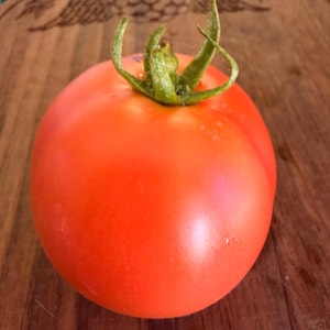 May include: A ripe, red tomato with a green stem sits on a wooden cutting board. The tomato is round and smooth, with a vibrant color. The cutting board has a dark wood grain and an engraved design.