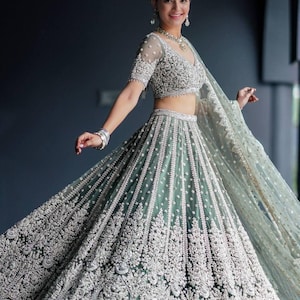 May include: A woman wearing a teal and silver embroidered lehenga with a matching dupatta. The lehenga has a full skirt and a fitted bodice. The dupatta is draped over the woman's head and shoulders.