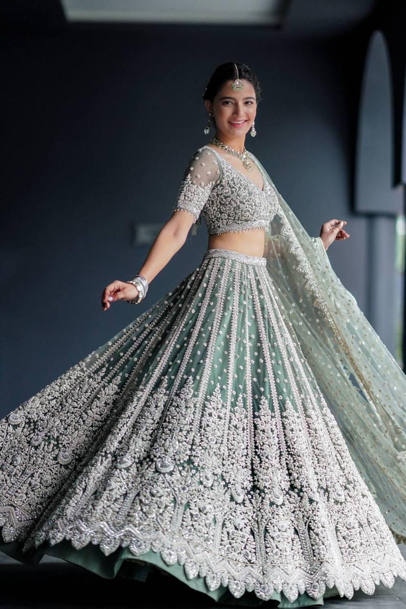 May include: A woman wearing a teal and silver embroidered lehenga with a matching dupatta. The lehenga has a full skirt and a fitted bodice. The dupatta is draped over the woman's head and shoulders.