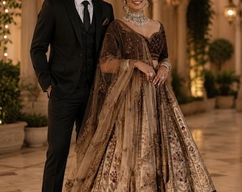 Può includere: Un abito nero a tre pezzi con camicia bianca e cravatta nera abbinato a un lehenga marrone scuro e crema con intricati ricami floreali, un ampio bordo scuro e una dupatta trasparente coordinata drappeggiata sulle spalle e braccia.