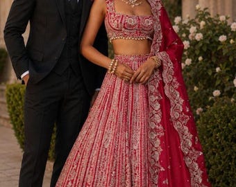 May include: A black three-piece suit with a white shirt and black tie, and a deep red lehenga set featuring a sleeveless crop top and a voluminous floor-length skirt, both heavily embellished with intricate silver embroidery and beadwork.