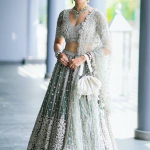 May include: A woman wearing a silver and green embroidered lehenga with a matching dupatta. The lehenga has a deep V-neckline and is embellished with sequins and beads. The dupatta is sheer and has a delicate floral pattern.