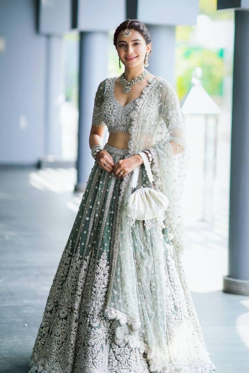 May include: A woman wearing a silver and green embroidered lehenga with a matching dupatta. The lehenga has a deep V-neckline and is embellished with sequins and beads. The dupatta is sheer and has a delicate floral pattern.