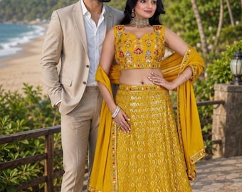 Könnte beinhalten: Ein beigefarbener Anzug mit weißem Hemd und braunen Slippern, kombiniert mit einem senfgelben Lehenga Choli mit aufwendiger Blumenstickerei, passendem Dupatta und einem weiten Rock mit detaillierten Verzierungen und Bogensaum.
