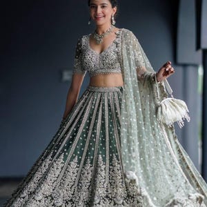 May include: A woman wearing a green and silver embroidered lehenga with a matching choli and dupatta. She is holding a white clutch purse.