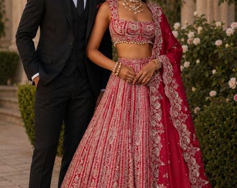 May include: A black three-piece suit with a white shirt and black tie, and a red lehenga choli featuring intricate silver embroidery, a sleeveless cropped bodice, a high-waisted pleated skirt, and a matching red dupatta with a wide silver border.