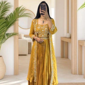 May include: Mustard yellow three-piece outfit. The set includes a crop top with floral embroidery, a long skirt, and a long-sleeved jacket with silver embellishments. The outfit is worn by a person in a room with a mirror and plants.