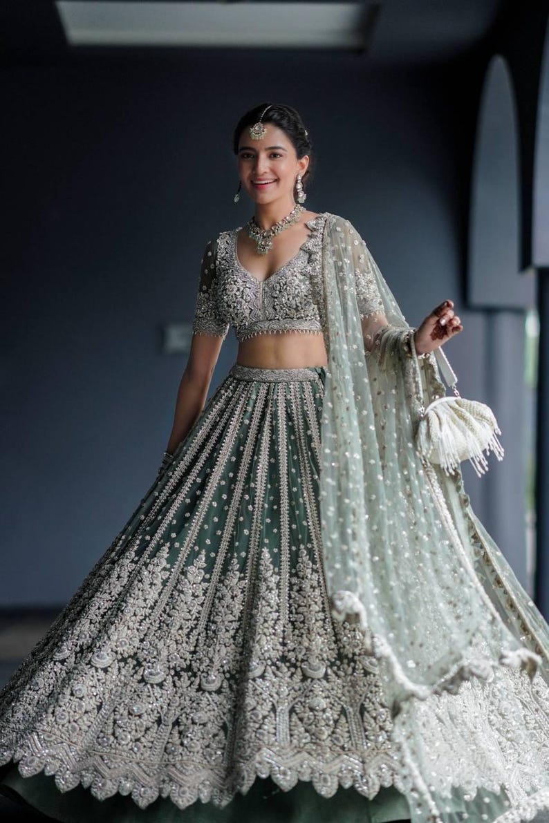 May include: A woman wearing a green and silver embroidered lehenga with a matching choli and dupatta. She is holding a white clutch purse.