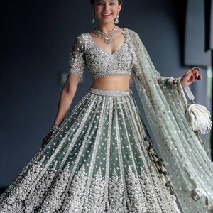 May include: A woman wearing a green and silver embroidered lehenga with a matching dupatta. The lehenga has a fitted bodice and a flowing skirt. The dupatta is draped over her shoulders and head.