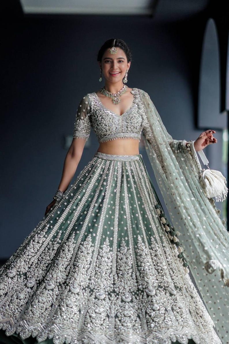May include: A woman wearing a green and silver embroidered lehenga with a matching dupatta. The lehenga has a fitted bodice and a flowing skirt. The dupatta is draped over her shoulders and head.
