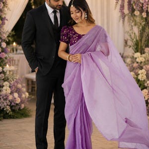 May include: A black three-piece suit with a white shirt and black tie worn alongside a lavender saree featuring a deep purple blouse with gold embroidery, a gold maang tikka headpiece, and gold bangles, showcasing a formal ensemble.
