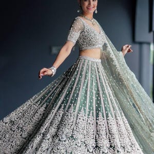 May include: A woman wearing a teal and silver embroidered lehenga with a matching dupatta. The lehenga has a full skirt and a fitted bodice. The dupatta is draped over the woman's head and shoulders.