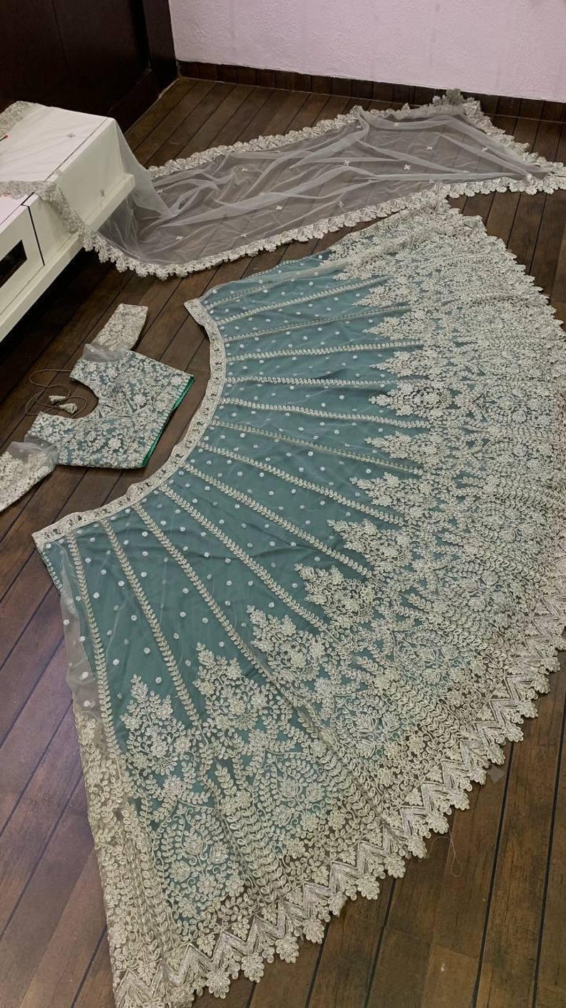 May include: A teal net lehenga with silver embroidery and a matching embroidered blouse. The lehenga has a scalloped hem and the blouse has a round neckline.