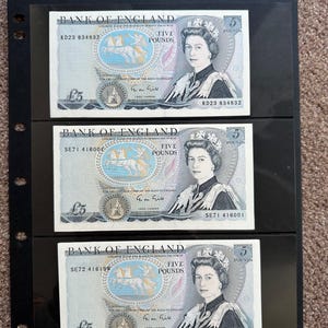 May include: Three Bank of England five-pound notes displayed in a black holder. Each note features a portrait of Queen Elizabeth II and a classical scene with horses. The notes are in good condition and are a collectible item.