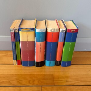 Può includere: Una collezione di sette libri di Harry Potter di J.K. Rowling disposti in fila. I libri presentano dorsi colorati con titoli che includono The Philosopher's Stone, Chamber of Secrets, Order of the Phoenix, Deathly Hallows, Goblet of Fire e Half-Blood Prince.