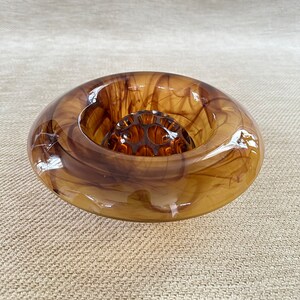 May include: A round, amber-colored glass flower frog with a dark brown, swirled pattern. The frog has a circular shape with a central grid of holes for arranging flowers. The glass has a glossy finish.