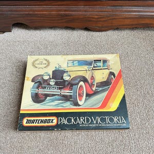 Può includere: Scatola del modellino Matchbox Packard Victoria. La scatola presenta un'illustrazione di un'auto d'epoca gialla e marrone con ruote rosse. La scatola riporta la scritta "PACKARD VICTORIA" e "MODELKIT-MAQUETTE-MODELL-BAUSATZ-BOUWMODEL".