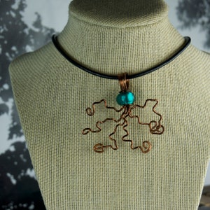 May include: A copper wire necklace with a teal blue bead pendant shaped like a tree. The necklace is on a black cord and is displayed on a tan fabric mannequin.