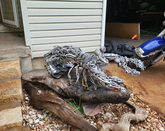 May include: A large metal sculpture of a lobster, crafted from recycled metal parts, sits on a weathered piece of driftwood. The sculpture is in a garden with gravel and a house in the background.