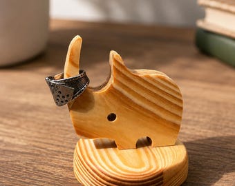 Op de afbeelding: Een lichtbruin houten neushoornbeeldje met een gladde, gepolijste afwerking en zichtbare houtnerf, gemonteerd op een ronde houten voet. Een zilverkleurige metalen ring met een getextureerd, geperforeerd patroon zit om de hoorn van de neushoorn.