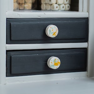 Könnte beinhalten: Zwei schwarze Schubladen mit weißen Verzierungen und dekorativen Knöpfen. Die Knöpfe sind rund und haben einen weißen Hintergrund mit einem Terrazzo-Muster in Gelb, Blau und Grün. Die Schubladen sind Teil eines weißen Schranks mit einer Glastür.