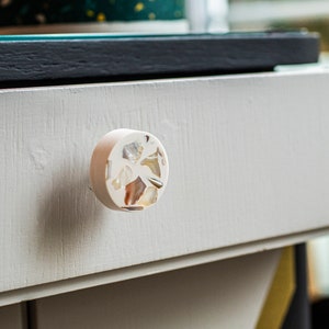 Könnte beinhalten: Ein runder, weißer Schubladengriff mit einem dekorativen Design aus eingebetteten Muscheln. Der Griff ist aus einem glatten, weißen Material gefertigt und hat einen leicht erhöhten Rand.