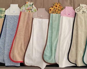 May include: A collection of hanging kitchen towels in various colors and patterns. Designs include floral, plaid, and solid colors, with contrasting trim. Each towel has a button closure at the top for hanging. The towels are made of soft, absorbent fabric.