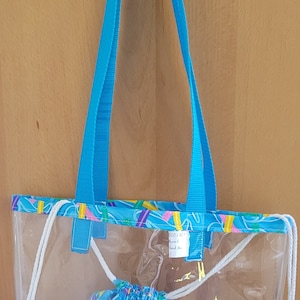 May include: Clear tote bag with blue handles and a matching drawstring bag. The tote features a colorful patterned trim and a smaller drawstring bag with a similar design. The bag is hanging on a white hook.
