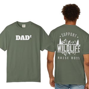 May include: An olive green short-sleeve t-shirt featuring the white text DAD² on the front. The back displays a white graphic of mountains and pine trees with the text SUPPORT WILDLIFE RAISE BOYS, framed by two stars at the top.