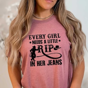 May include: A heather mauve t-shirt with the text "Every girl needs a little rip in her jeans" in black. The text is accompanied by a silhouette of a cowgirl roping a calf.