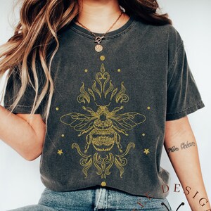 May include: Pepper-colored t-shirt featuring a detailed gold-colored bee design with ornate embellishments. The design includes a bee, decorative flourishes, and stars. The shirt is paired with blue jeans.