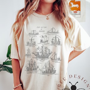 May include: A beige t-shirt with a black and white illustration of different types of ships. The text "Some old types of ships" is printed above the illustration. The t-shirt has a crew neck and short sleeves. The t-shirt is on a model wearing black pants.
