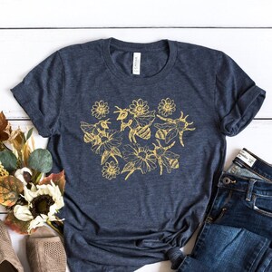 May include: A navy blue t-shirt with a gold floral and bee design. The shirt is paired with blue jeans and tan open-toe shoes. A bouquet of fall-colored flowers and foliage is arranged to the left of the shirt.