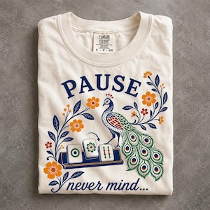 May include: An off-white short-sleeve t-shirt featuring a graphic of a peacock standing next to three mahjong tiles. The text reads PAUSE above the bird and never mind... below. The design includes orange flowers and dark blue leafy vines.