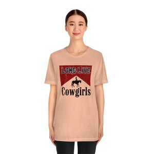 May include: A peach-colored t-shirt featuring a distressed graphic. The graphic includes the text "LONG LIVE" above a silhouette of a person riding a horse, with "Cowgirls" below. The shirt has short sleeves and a crew neck.
