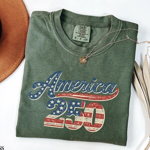 Pode incluir: Uma camiseta verde musgo com o texto "America 250" em um design vintage e patri&oacute;tico. O texto apresenta um esquema de cores vermelho e azul, com estrelas e listras incorporadas ao design. Um colar de ouro repousa sobre a camisa.