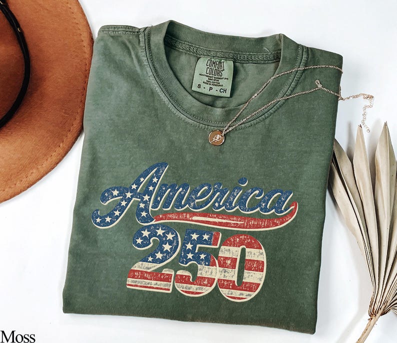 Pode incluir: Uma camiseta verde musgo com o texto "America 250" em um design vintage e patri&oacute;tico. O texto apresenta um esquema de cores vermelho e azul, com estrelas e listras incorporadas ao design. Um colar de ouro repousa sobre a camisa.