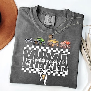 May include: Dark gray t-shirt with a graphic of monster trucks and the word "MAMA" in white block letters. A checkered flag and a trophy are also included in the design. A silver necklace with a pendant is draped across the shirt.