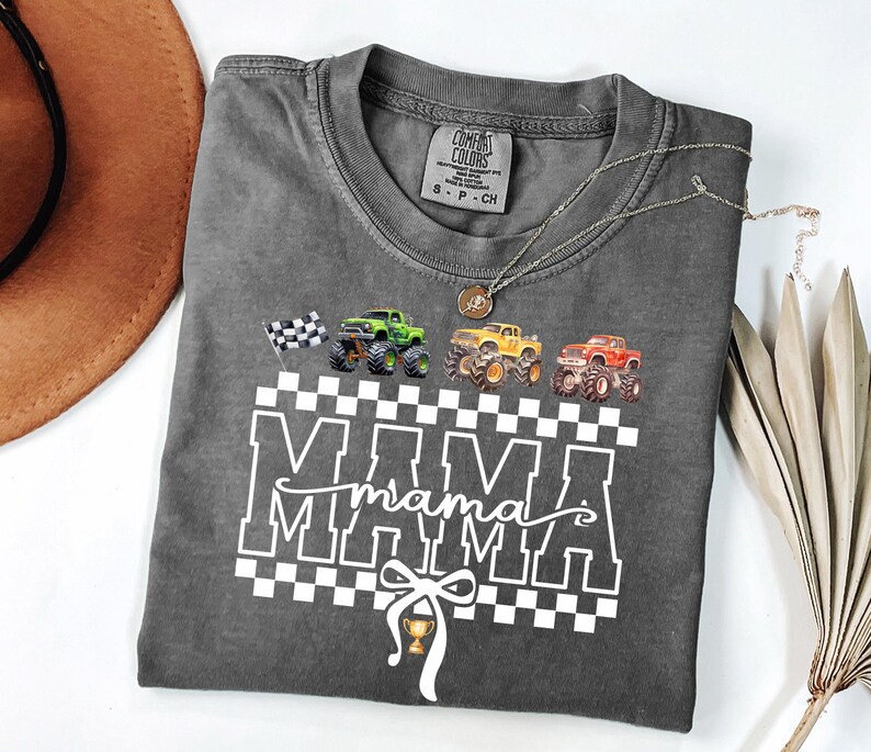 May include: Dark gray t-shirt with a graphic of monster trucks and the word "MAMA" in white block letters. A checkered flag and a trophy are also included in the design. A silver necklace with a pendant is draped across the shirt.