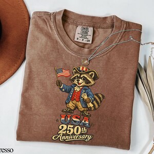 Comfort Colors 1717 garment-dyed t-shirt in Espresso brown featuring a vintage-style 'USA 250th Anniversary' graphic. The illustration depicts a patriotic raccoon 'trash panda' dressed as a Founding Father in Revolutionary War attire holding an American flag. Perfect retro Semiquincentennial gift for history lovers celebrating America's 250th birthday.